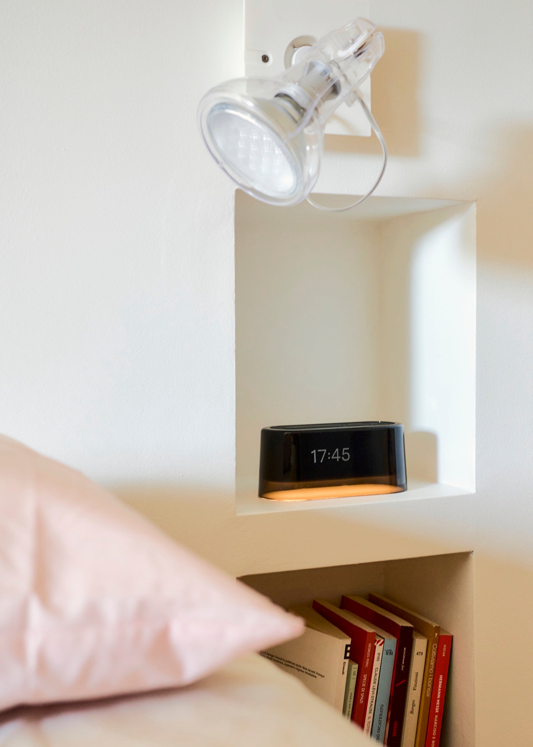A black Loftie Clock glowing in a white wall nook above a bookshelf, beside a bed. Photo by Alecio Ferrari (@alecio.untitled).